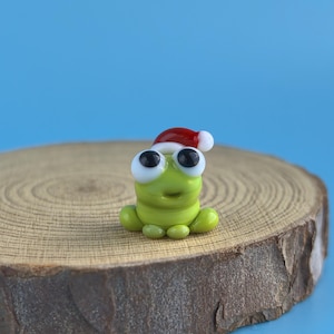 May include: A small, green frog figurine with large, black-eyed eyes and a smiling expression. The frog is wearing a red and white Santa hat. It is sitting on a round, wooden slice against a blue background.