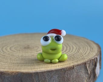 May include: A small, green frog figurine with large, black-eyed eyes and a smiling expression. The frog is wearing a red and white Santa hat. It is sitting on a round, wooden slice against a blue background.