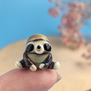 May include: A miniature glass sloth figurine with a striped head, large black eyes, and a light-colored belly. The sloth is sitting with its arms and legs outstretched. The figurine is primarily brown, black, and white, with a touch of yellow.