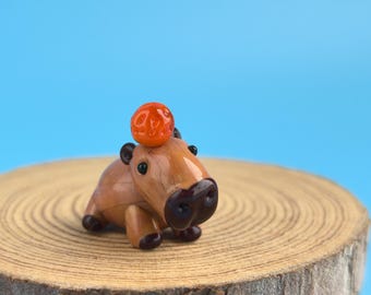 May include: A miniature, handcrafted capybara figurine in brown, with a small orange sphere on its head. The figurine is posed on a wooden slice against a light blue backdrop, highlighting the animal's features.