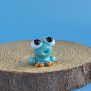 May include: A small, turquoise glass frog figurine with large, black-pupiled eyes and orange feet. The frog is perched on a round, wooden slice against a solid blue background. The figurine is a whimsical decorative item.