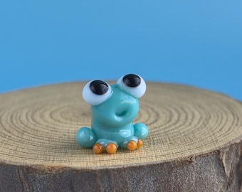 May include: A small, turquoise glass frog figurine with large, black-pupiled eyes and orange feet. The frog is perched on a round, wooden slice against a solid blue background. The figurine is a whimsical decorative item.