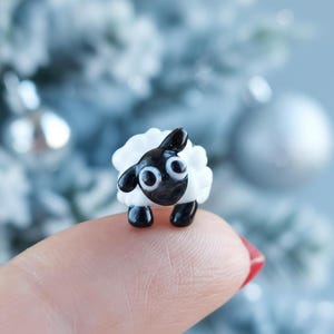 May include: A miniature glass sheep figurine with a black head and legs, and a fluffy white body. The sheep has large, round eyes and is resting on a fingertip. The background is blurred with silver ornaments.