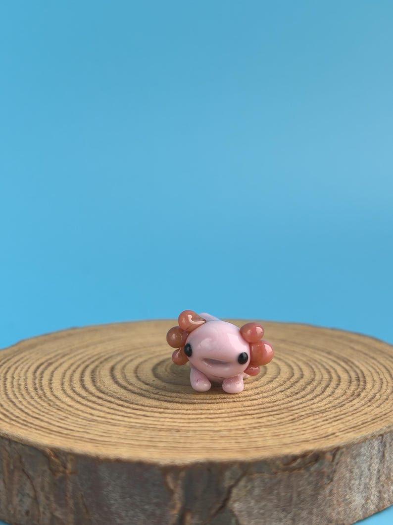 Op de afbeelding: Een kleine, roze axolotl-figuur met oranje kieuwen, geplaatst op een houten schijf tegen een blauwe achtergrond. De axolotl heeft kleine pootjes en zwarte ogen. Het beeldje heeft een glimlachende uitdrukking.