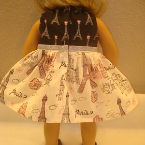 May include: A doll wearing a dress with a black bodice featuring Eiffel Tower designs. The skirt is white with pink and brown Parisian illustrations and the word "Paris". The dress has a silver glitter trim.