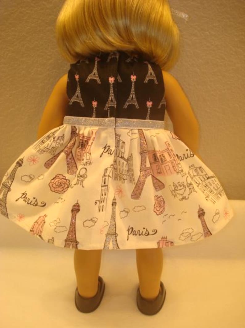 May include: A doll wearing a dress with a black bodice featuring Eiffel Tower designs. The skirt is white with pink and brown Parisian illustrations and the word "Paris". The dress has a silver glitter trim.