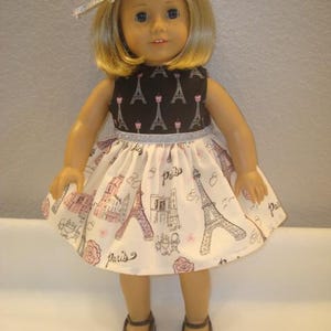 May include: A doll wearing a black and white dress with a Paris theme. The dress features a black bodice with Eiffel Tower prints and a white skirt with pink and black Parisian illustrations. The doll has blonde hair and brown shoes.