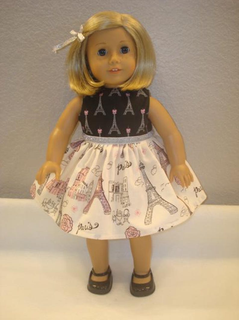 May include: A doll wearing a black and white dress with a Paris theme. The dress features a black bodice with Eiffel Tower prints and a white skirt with pink and black Parisian illustrations. The doll has blonde hair and brown shoes.