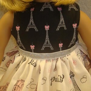 May include: A doll dress featuring a black bodice with Eiffel Tower and pink bow prints. The skirt is white with a Paris theme, including the Eiffel Tower and the word "Paris". A silver ribbon accents the waist.