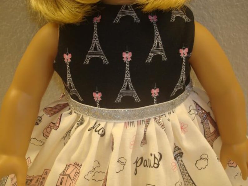 May include: A doll dress featuring a black bodice with Eiffel Tower and pink bow prints. The skirt is white with a Paris theme, including the Eiffel Tower and the word "Paris". A silver ribbon accents the waist.