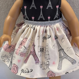 May include: A doll dress featuring a black bodice with Eiffel Tower and pink bow accents. The skirt is cream-colored with a Paris theme, including illustrations of the Eiffel Tower, buildings, and the word "Paris". A silver glitter band accents the waist.