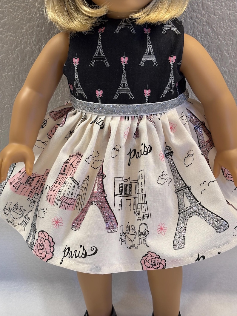 May include: A doll dress featuring a black bodice with Eiffel Tower and pink bow accents. The skirt is cream-colored with a Paris theme, including illustrations of the Eiffel Tower, buildings, and the word "Paris". A silver glitter band accents the waist.