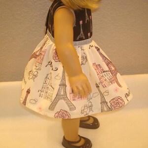 May include: A doll wearing a sleeveless dress with a black bodice and a white skirt. The skirt features a repeating pattern of the Eiffel Tower, buildings, and roses in pink and brown. The doll is wearing brown shoes.