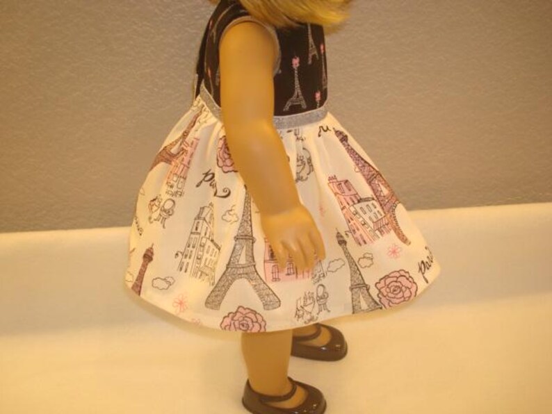 May include: A doll wearing a sleeveless dress with a black bodice and a white skirt. The skirt features a repeating pattern of the Eiffel Tower, buildings, and roses in pink and brown. The doll is wearing brown shoes.