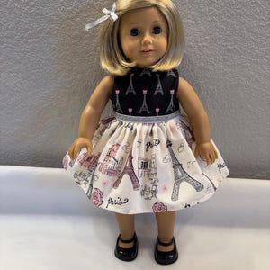 May include: A doll wearing a black and white dress with a Paris theme. The dress features a black bodice with Eiffel Tower designs and a white skirt with pink and black Parisian illustrations. The doll has blonde hair and black shoes.