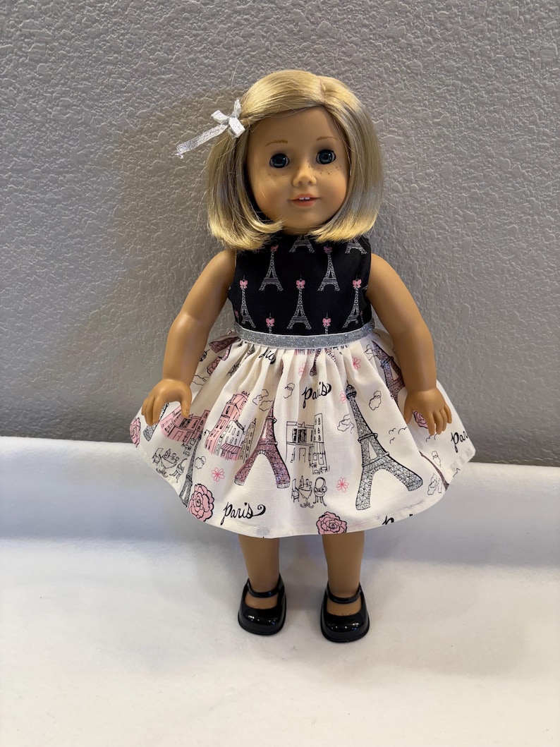 May include: A doll wearing a black and white dress with a Paris theme. The dress features a black bodice with Eiffel Tower designs and a white skirt with pink and black Parisian illustrations. The doll has blonde hair and black shoes.