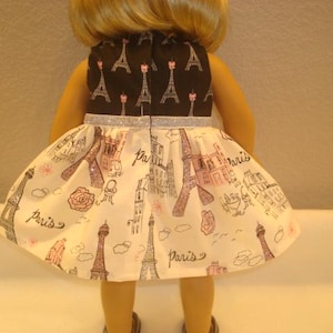May include: A doll wearing a dress with a black top featuring Eiffel Tower designs and a silver glitter belt. The skirt is white with illustrations of the Eiffel Tower, buildings, and the word "Paris".