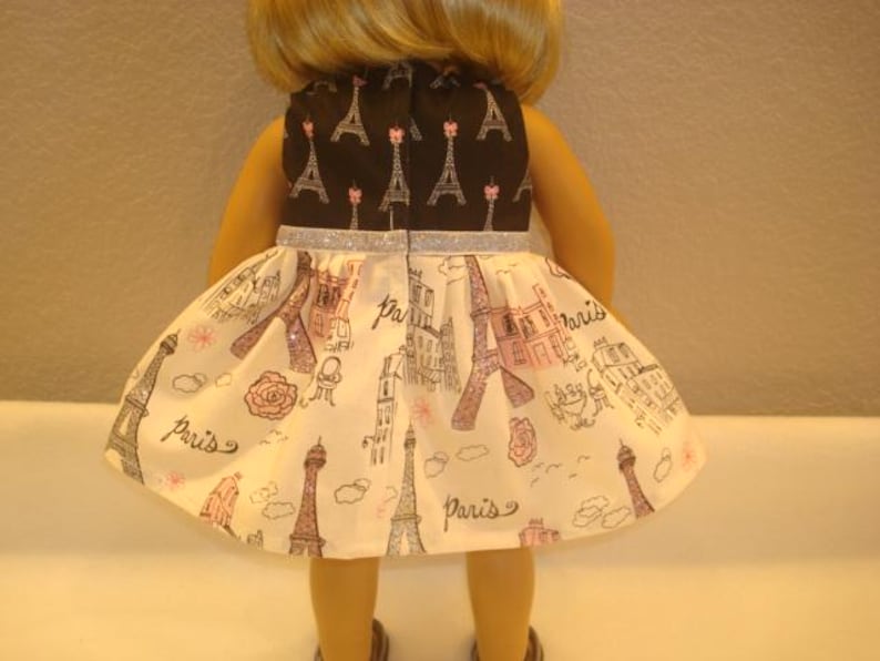 May include: A doll wearing a dress with a black top featuring Eiffel Tower designs and a silver glitter belt. The skirt is white with illustrations of the Eiffel Tower, buildings, and the word "Paris".