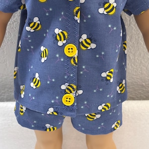 May include: Blue pajama set with a repeating pattern of yellow and black bees. The short-sleeved top has yellow buttons and a zig-zag stitched seam. The matching shorts complete the set. This sleepwear is designed for a doll.