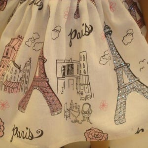 May include: A white doll dress with a Paris theme. The fabric features illustrations of the Eiffel Tower, buildings, and the word "Paris" in black script. The Eiffel Tower is accented with glitter.