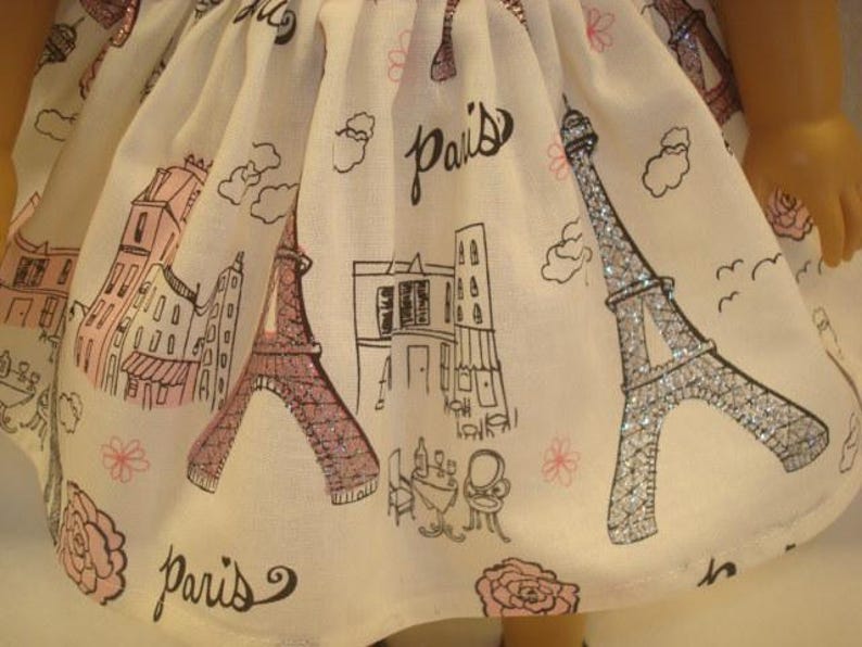 May include: A white doll dress with a Paris theme. The fabric features illustrations of the Eiffel Tower, buildings, and the word "Paris" in black script. The Eiffel Tower is accented with glitter.