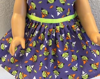May include: A sleeveless doll dress in a deep purple fabric featuring a repeating pattern of green witch faces with orange or white hats, interspersed with small orange candies. The waist is accented with a bright lime green ribbon trim.