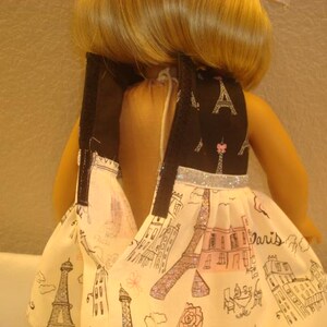 May include: A doll wearing a dress with a black bodice featuring Eiffel Tower and pink accents. The skirt is white with a Paris-themed print, including the Eiffel Tower and other landmarks. The dress has a silver glitter trim.