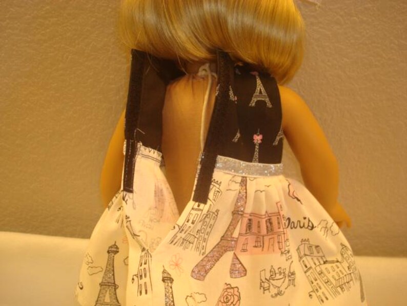 May include: A doll wearing a dress with a black bodice featuring Eiffel Tower and pink accents. The skirt is white with a Paris-themed print, including the Eiffel Tower and other landmarks. The dress has a silver glitter trim.