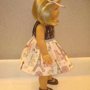 May include: A doll wearing a brown and white dress with a Parisian theme. The dress features illustrations of the Eiffel Tower, buildings, and roses. The doll has blonde hair and is wearing brown shoes.