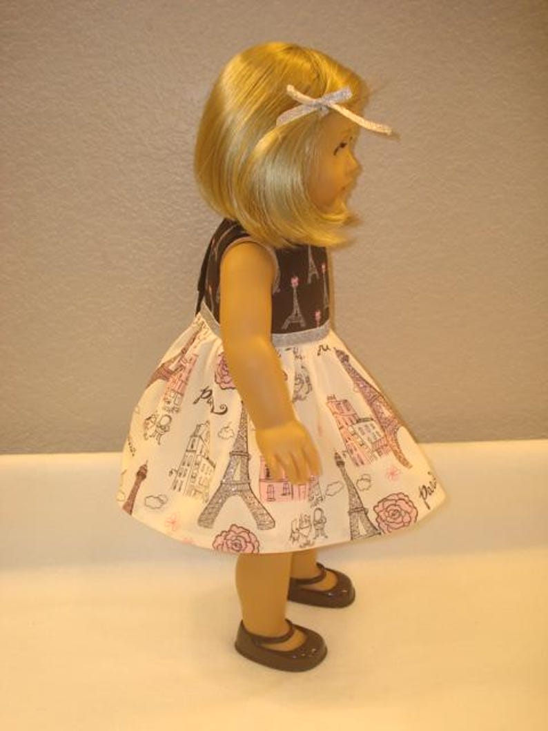May include: A doll wearing a brown and white dress with a Parisian theme. The dress features illustrations of the Eiffel Tower, buildings, and roses. The doll has blonde hair and is wearing brown shoes.