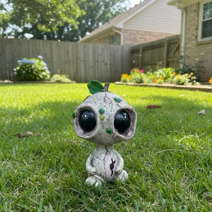 Puede incluir: Una figura caprichosa y desgastada con grandes ojos negros y una pequeña hoja verde en la parte superior. La figura tiene un cuerpo gris claro con acentos verdes y rosas, y está sentada sobre hierba verde.
