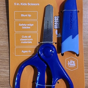 May include: Blue Fiskars 5-inch kids scissors with a blunt tip and safety-edge blades. The scissors come with a blue eraser sheath and are designed for classroom materials. The packaging includes the text "World's #1 Scissors Brand" and "Ages 4+".