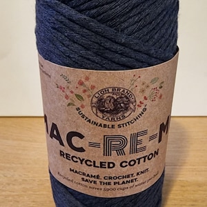 May include: A large ball of dark blue recycled cotton yarn. The label reads "Lion Brand Yarns" and "Sustainable Stitching." It is suitable for macrame, crochet, and knitting. The label also states that the recycled cotton saves 5900 cups of water per ball.