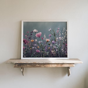 May include: A framed print depicting a wildflower meadow in hues of pink, purple, white, and yellow, set against a muted teal backdrop. The artwork is presented in a white frame and sits on a rustic wooden shelf.