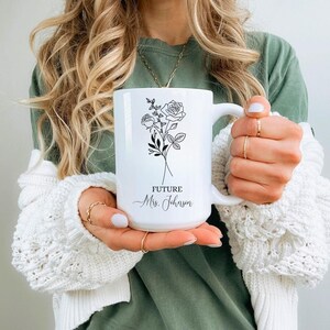 以下が含まれることがあります： 黒い花の模様（バラを含む）と「FUTURE Mrs. Johnson」の文字が入った白いセラミックマグ。マグには大きなハンドルが付いています。マグを持っている人は、グリーンのシャツと白いカーディガンを着ています。
