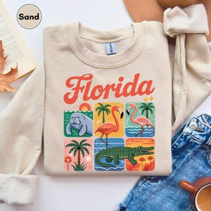 May include: Sand-colored sweatshirt with the word "Florida" in orange and white lettering. The design features colorful squares with illustrations of a manatee, flamingos, a palm tree, and an alligator.