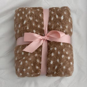Può includere: Una coperta rettangolare in pelliccia sintetica piegata, caratterizzata da una base marrone con un motivo a pois bianchi. La coperta è fissata con un nastro di raso rosa chiaro annodato al centro, creando una presentazione ordinata.