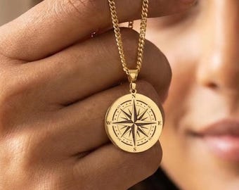 May include: Gold-colored compass pendant necklace. The pendant features a detailed compass rose design with directional markers. The necklace has a curb chain. The pendant is held up by a person's hand.