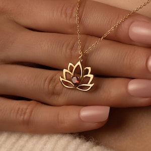Op de afbeelding: Een goudkleurige lotusbloem hanger ketting met een kleine, rode edelsteen in het midden. De ketting is delicaat, met een fijne ketting. Het lotusbloem ontwerp is open en gedetailleerd, met de edelsteen. Het is een sieraad.