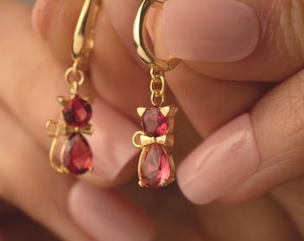 Op de afbeelding: Een paar gouden oorringen met een hangende rode kattenhanger van edelsteen. De oorringen zijn van het creolen-type met een klein kettinkje dat de creolen met de hanger verbindt.