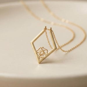 May include: A delicate gold necklace featuring a diamond-shaped pendant with a floral design at its center. The pendant is suspended from a fine gold chain, creating an elegant and minimalist aesthetic. The necklace is displayed on a white surface.