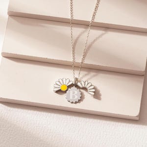 May include: A silver necklace with a charm featuring a daisy with a yellow center and the letter "B" in the center of the charm.