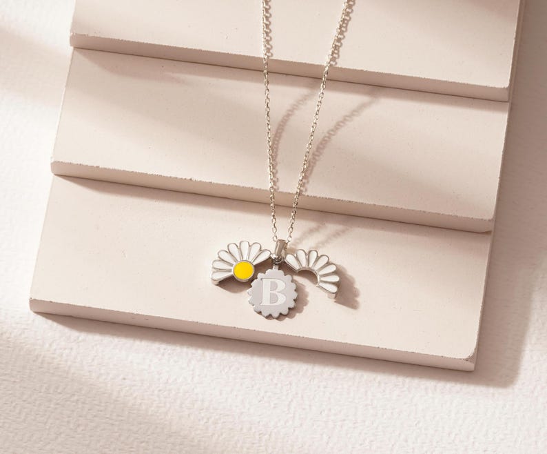 May include: A silver necklace with a charm featuring a daisy with a yellow center and the letter "B" in the center of the charm.