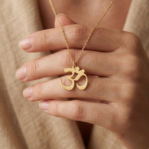 May include: A gold-colored necklace featuring the Om symbol pendant. The pendant is a stylized representation of the sacred sound. The necklace is displayed against a neutral background, highlighting the jewelry's design and color.