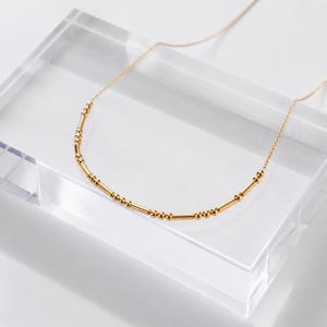 Könnte beinhalten: Eine goldfarbene Halskette mit einer feinen Kette und einem Mittelstück aus abwechselnden kleinen runden Perlen und kurzen zylindrischen Stäbchen, die in einem an Morsezeichen erinnernden Muster angeordnet und bogenförmig aufgehängt sind.