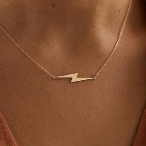 May include: A gold-colored necklace featuring a lightning bolt pendant. The pendant is a stylized zig-zag shape, suspended from a delicate chain. The necklace is worn around the neck, with the chain resting against the skin.