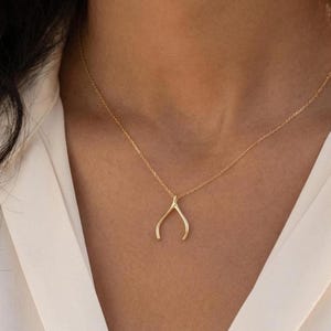 May include: A delicate gold-colored necklace featuring a wishbone pendant. The pendant hangs from a fine chain, creating a minimalist and elegant look. The necklace is worn against a white blouse.