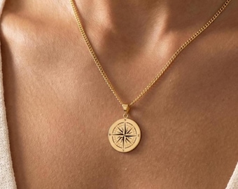 May include: A gold-colored necklace featuring a round pendant with a compass rose design. The pendant hangs from a delicate chain. The necklace is worn around the neck.