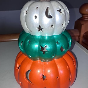 Peut inclure: Une décoration en forme de citrouilles empilées sur trois niveaux, de couleurs blanche, verte et orange. Chaque section présente une finition brillante et des découpes en forme d'étoiles et de croissants de lune, avec une tige brune au sommet.