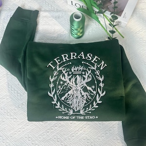 Może przedstawiać: Zielony sweter z białym haftem. Haft przedstawia jelenia z rogami, koronę i napis "TERRASEN" oraz "HOME OF THE STAG".